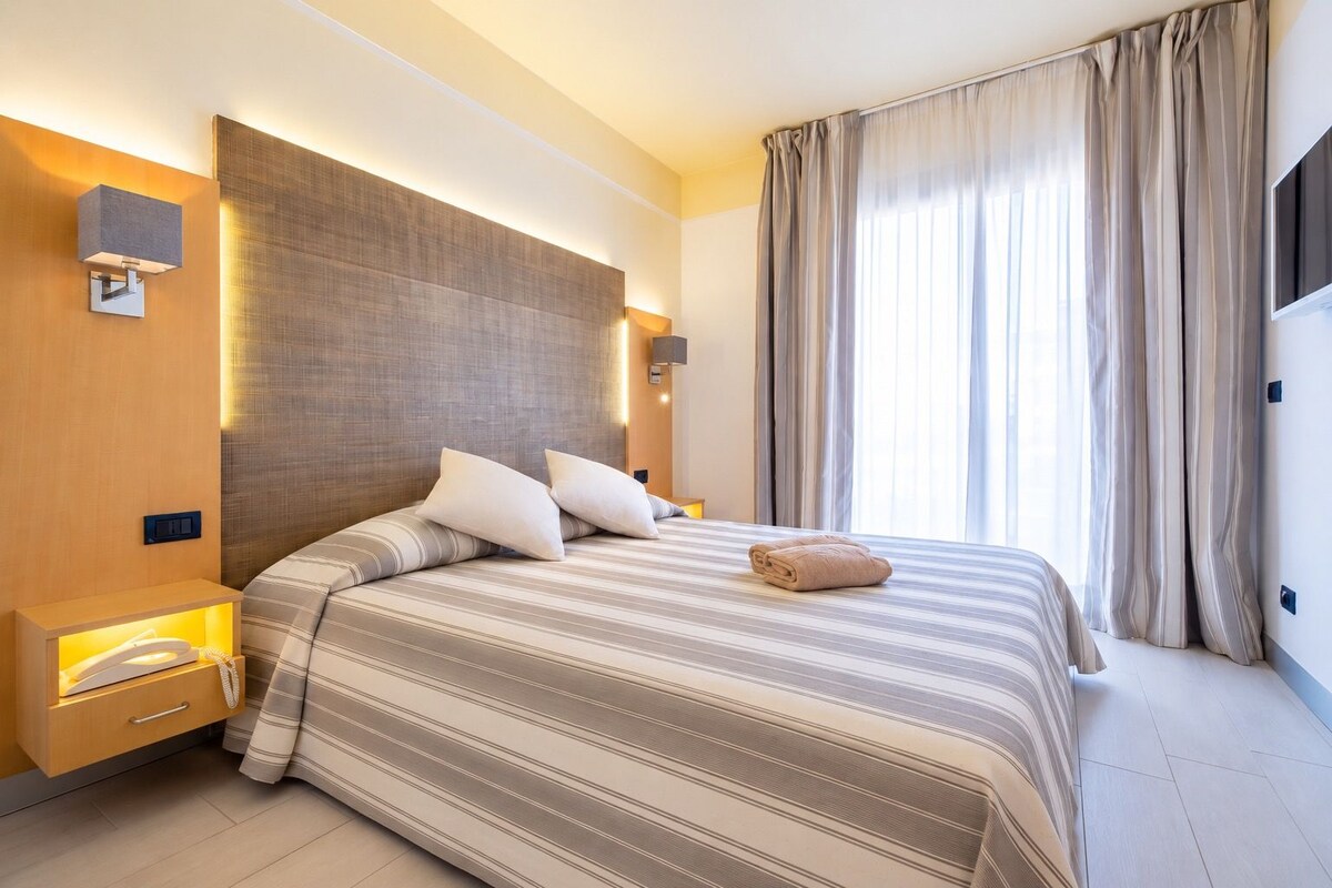 Una camera d'albergo con un letto grande, lampade sui comodini, una TV a schermo piatto e una finestra con tende.