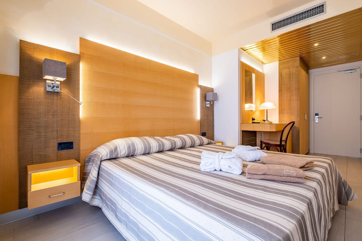 A modern hotel room with a large bed, a bedside table with a lamp, a mirror, and a small desk with a chair.