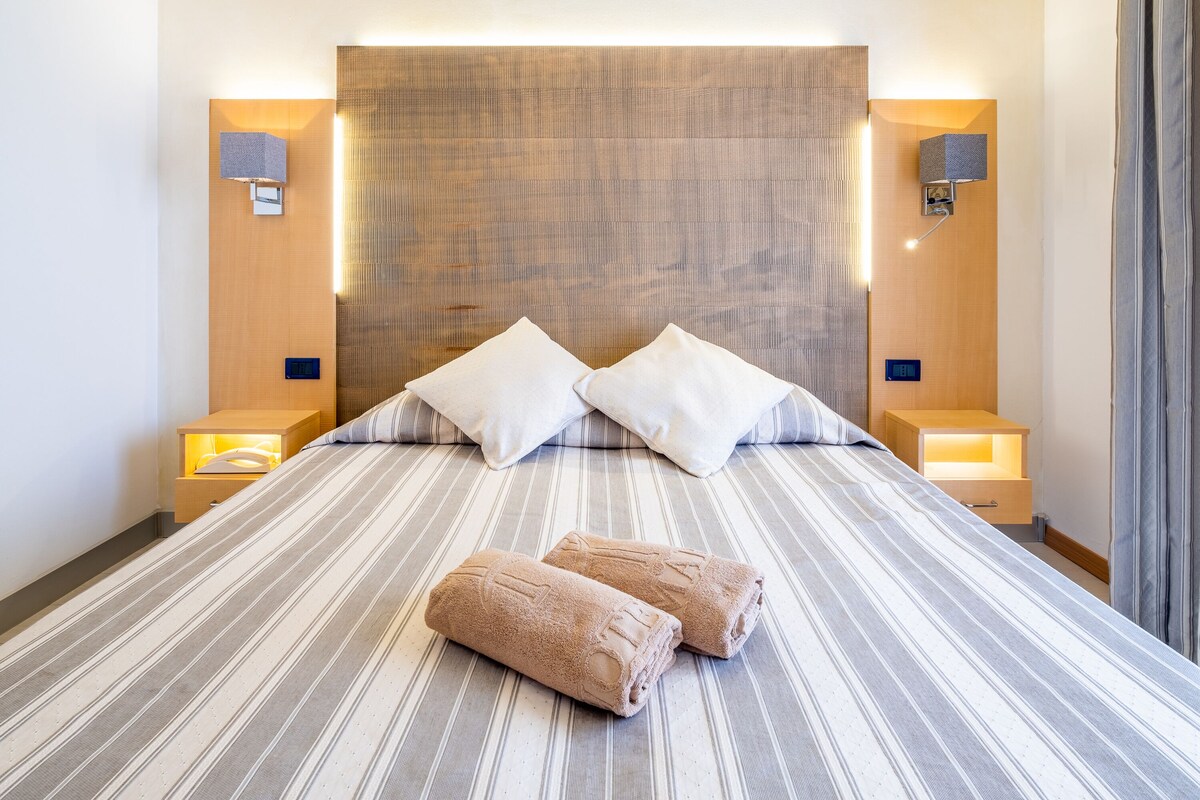 A hotel room with a large bed, two pillows, two bedside tables with lamps, and a headboard with a textured pattern.