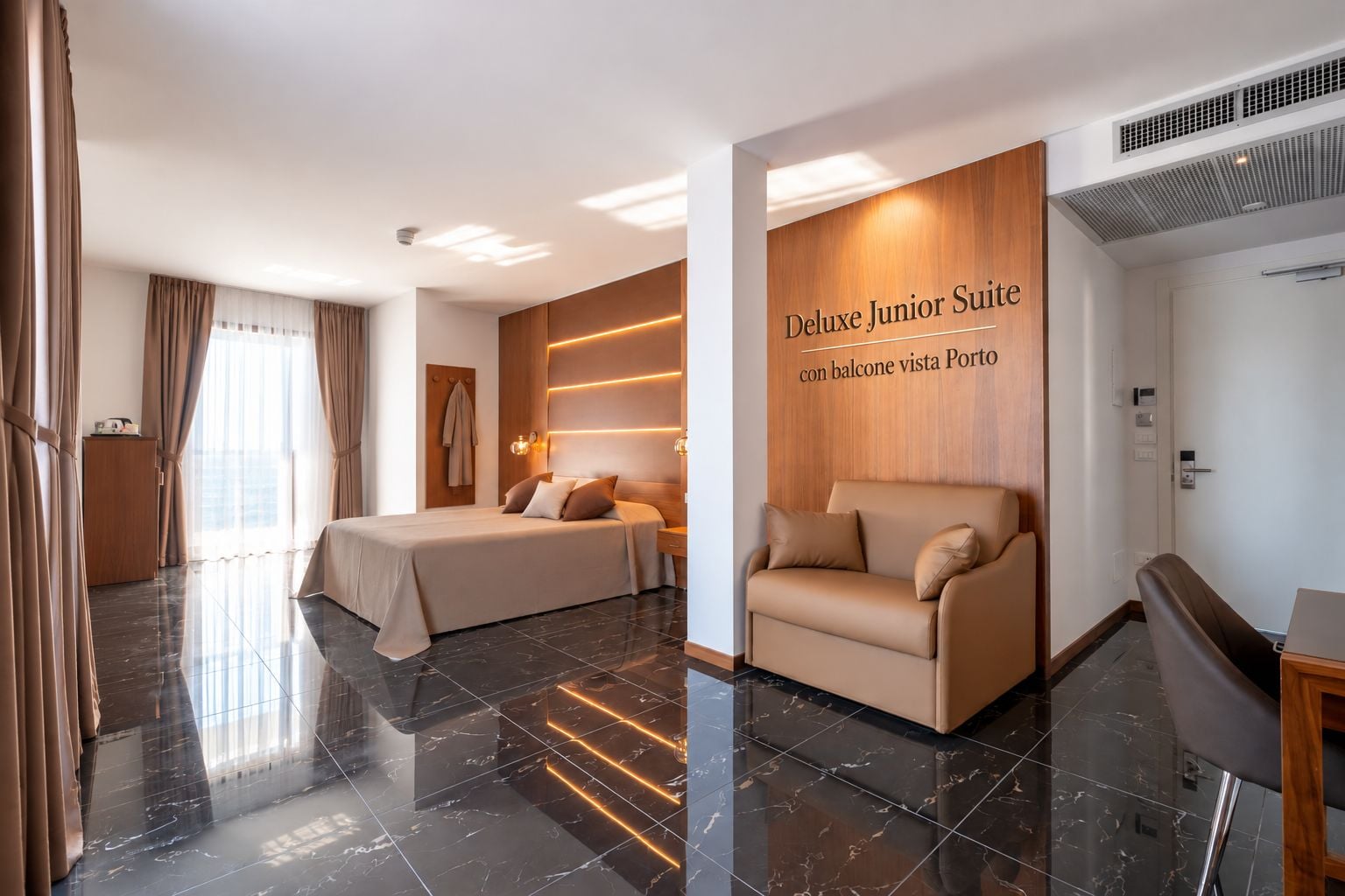 A Deluxe Junior Suite with a balcony, a bed, a sofa, and a desk.