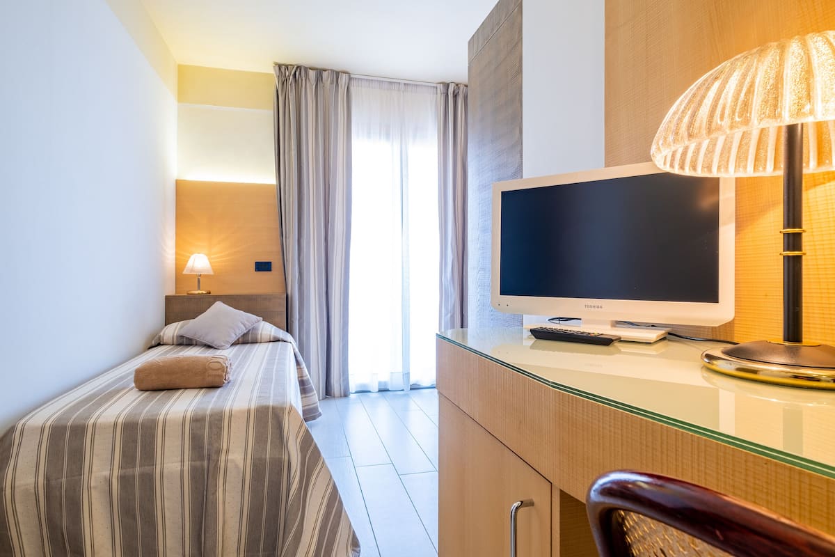 A hotel room with a bed, a desk with a computer, a lamp, and a window with curtains.