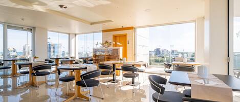 A modern dining area with multiple tables and chairs, large windows offering city views, and a central decorative piece.