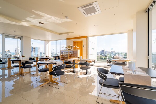 Zona pranzo moderna con diversi tavoli e sedie, ampie finestre con vista sulla città e un elemento decorativo centrale.