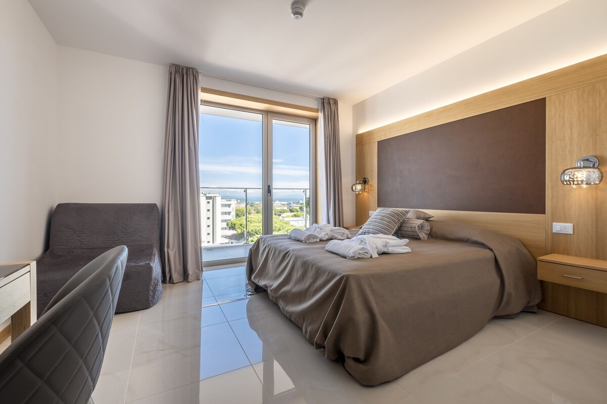 A modern hotel room with a large bed, a desk, a chair, and a view of the city through the window.