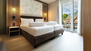 A modern hotel room with a large bed, a nightstand, a wall-mounted lamp, and a balcony with a view of the mountains. - Hotel Brione Green Resort (Riva del Garda)