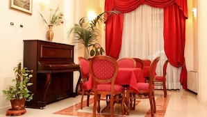 Un coin repas avec un piano, des chaises rouges, une table et une grande fenêtre ornée de rideaux rouges.