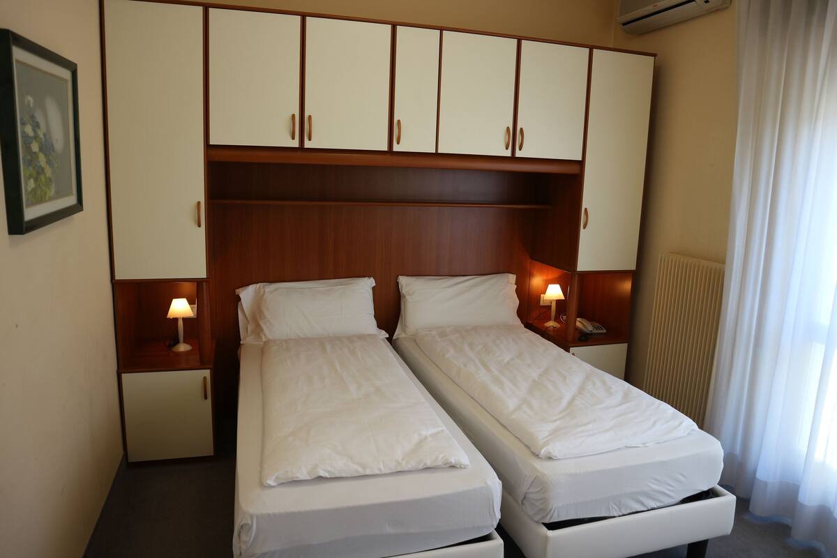 A hotel room with two single beds, wooden headboard, bedside lamps, a small cabinet, and a radiator.