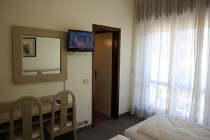 A hotel room with a bed, a desk with a chair, a TV mounted on the wall, and a door leading to another room. - Hotel Bristol (Riva del Garda)