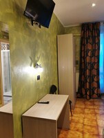 Una stanza con una scrivania, una sedia, un televisore fissato al muro e una tenda con un motivo a foglia.