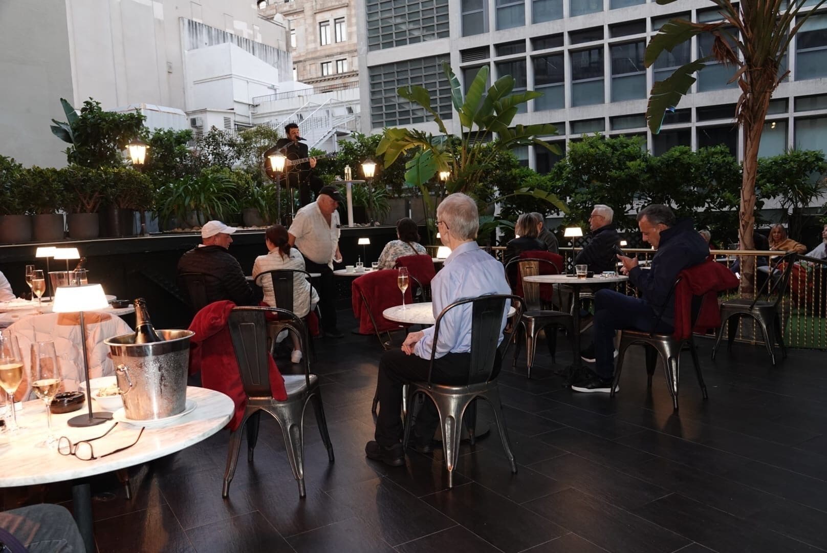 An outdoor dining area with tables and chairs, surrounded by greenery and buildings.