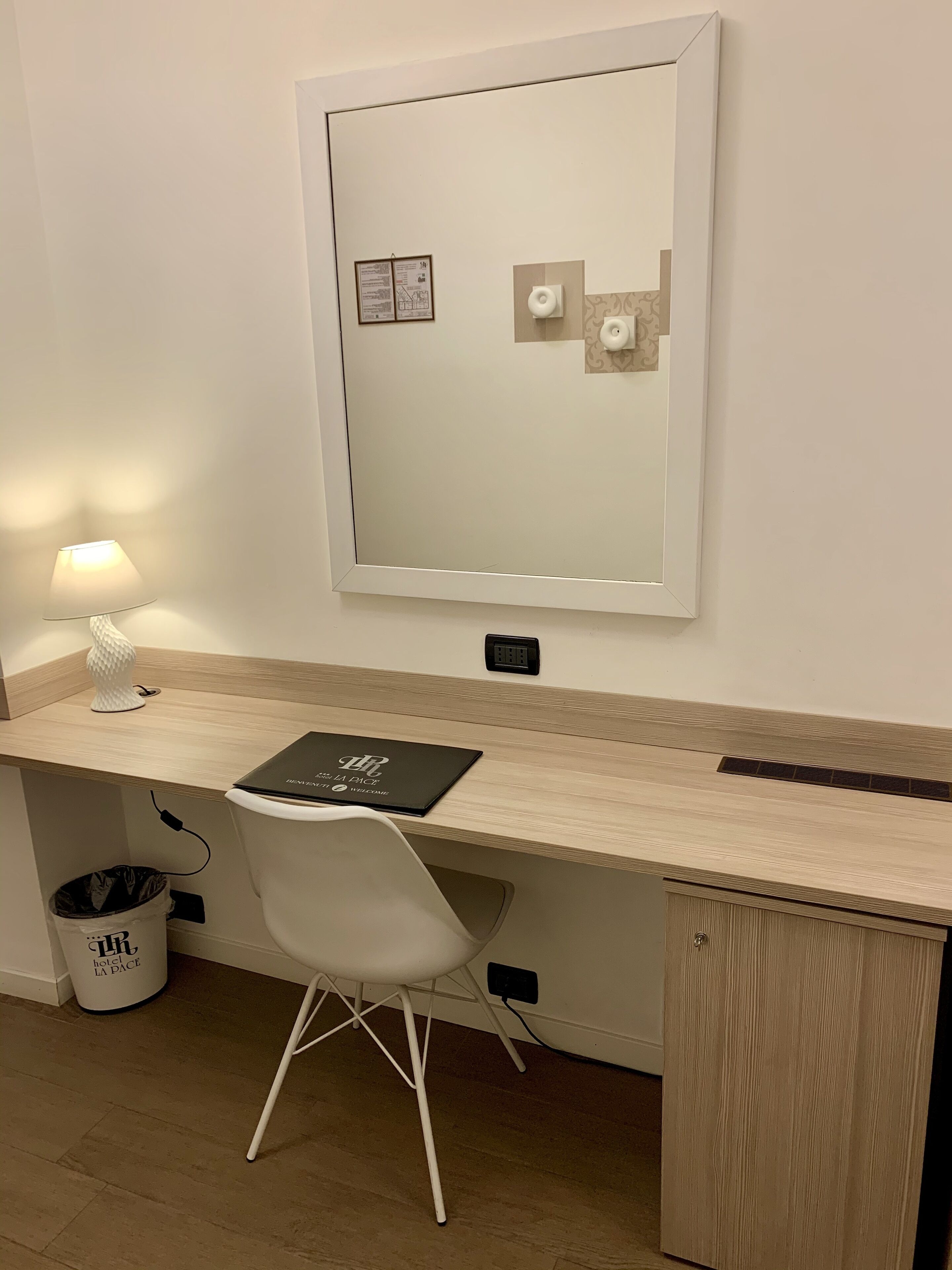 A hotel room with a wooden desk, a white chair, a small trash bin, a mirror, and a wall-mounted light.