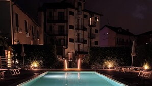 Una piscina rectangular con borde iluminado, rodeada de sillas de descanso y sombrillas, con edificios al fondo.