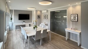 Un comedor moderno con una mesa larga, sillas, luces empotradas en el techo, un televisor de pantalla plana y un ventanal grande.