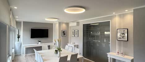 A modern dining area with a long table, chairs, recessed ceiling lights, a flat-screen TV, and a large window.