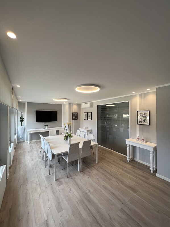 A modern dining area with a long table, chairs, recessed ceiling lights, a flat-screen TV, and a large window. - La Pace (San Mauro Torinese)