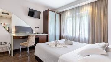 Una camera d'albergo con un letto, una scrivania, una sedia, un televisore a parete e un'ampia finestra con tende.