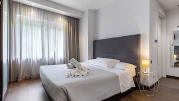 Habitación de hotel moderna con una cama grande, una mesita de noche con lámpara y vistas al exterior a través de cortinas translúcidas.