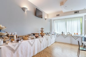 Disposição de buffet de café da manhã com diversos alimentos e bebidas, uma televisão presa na parede e uma grande janela com cortinas.
