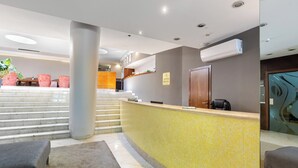 Ein Hotel-Empfangsbereich mit gelb gefliestem Empfangstresen, einem Schreibtisch, einer Treppe zu den oberen Etagen und einer Sitzecke mit gepolsterten Stühlen.