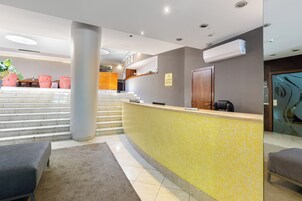 Uma área de recepção de hotel com balcão revestido de azulejos amarelos, um balcão de atendimento, uma escadaria que leva aos andares superiores e uma área de estar com cadeiras estofadas.