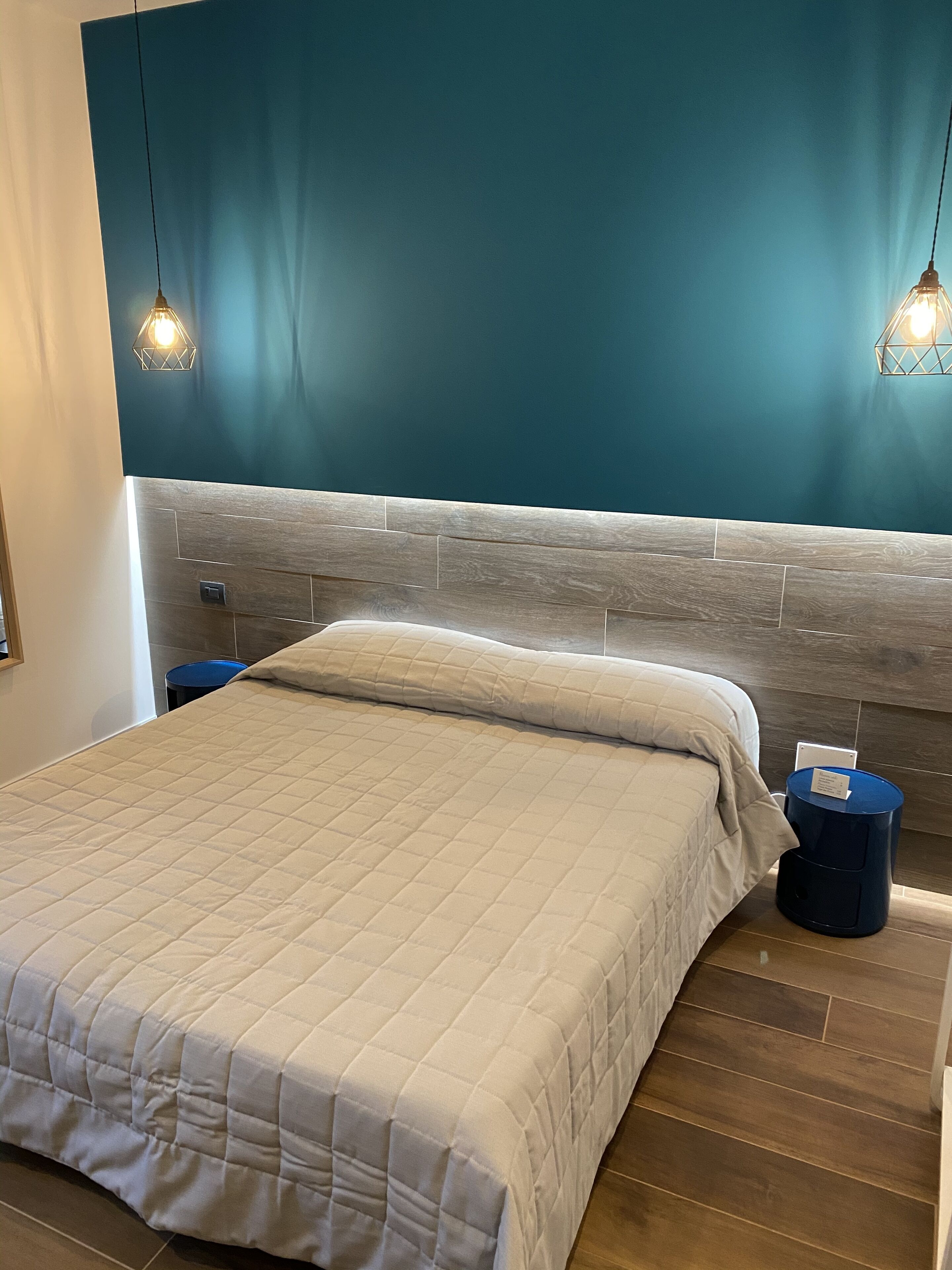 A modern hotel room with a large bed, a wooden headboard, teal walls, and two pendant lights.