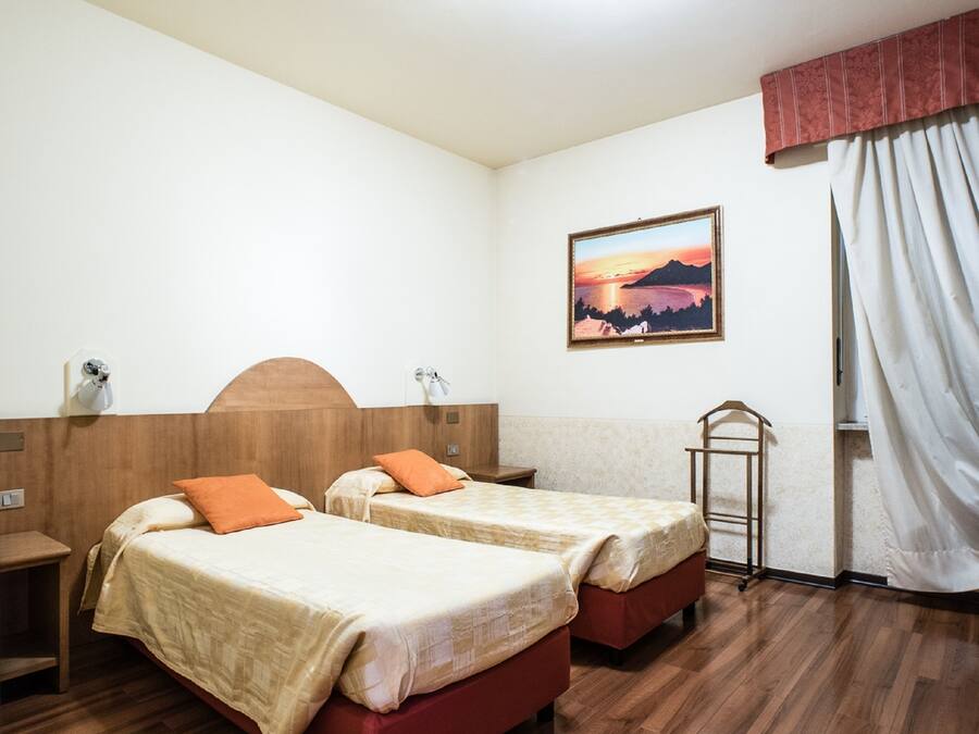 A hotel room with two beds, wooden flooring, a painting on the wall, and a window with curtains.
