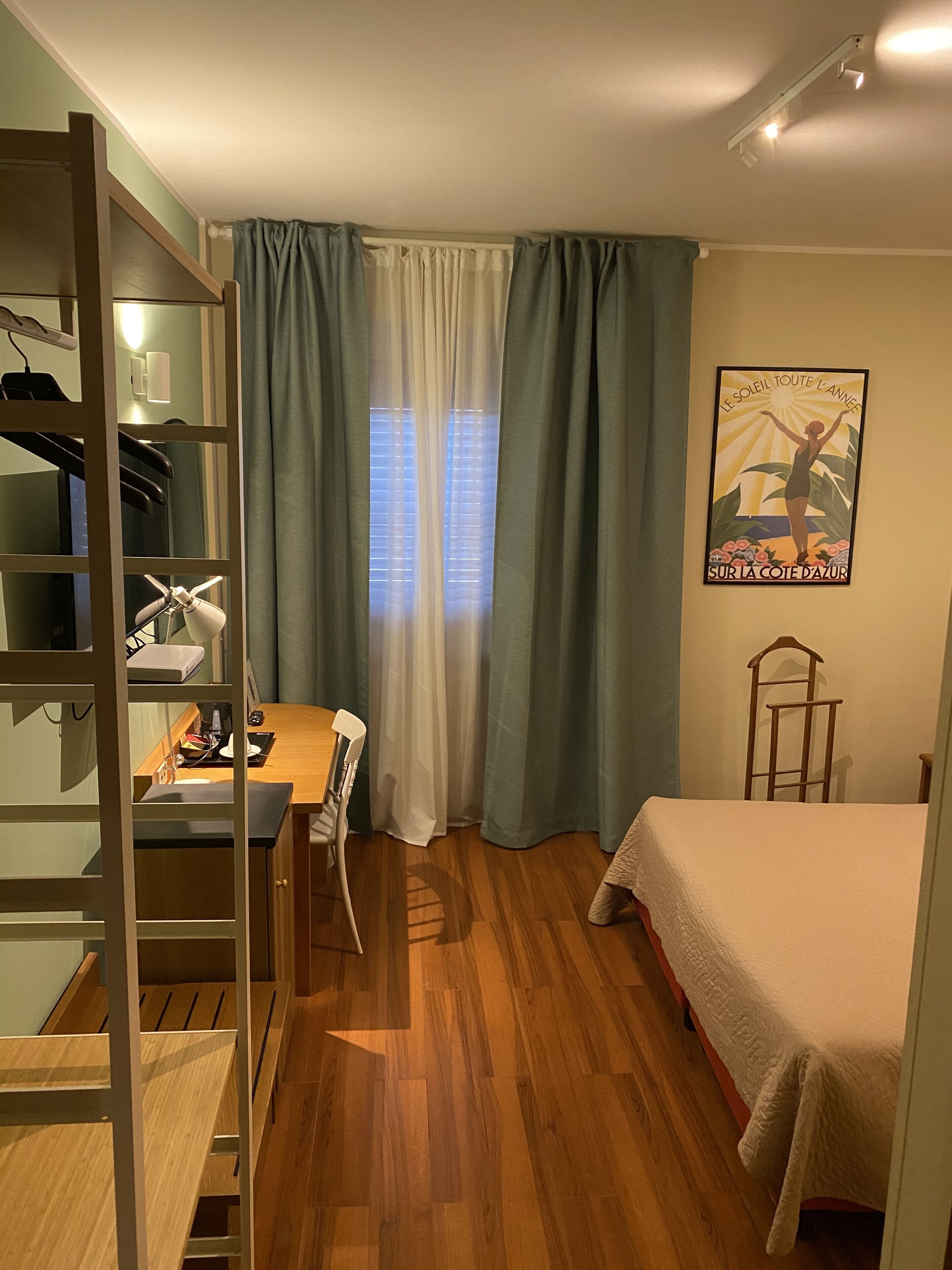 A bedroom with a wooden bed, a desk with a chair, a wardrobe, and a window with curtains.