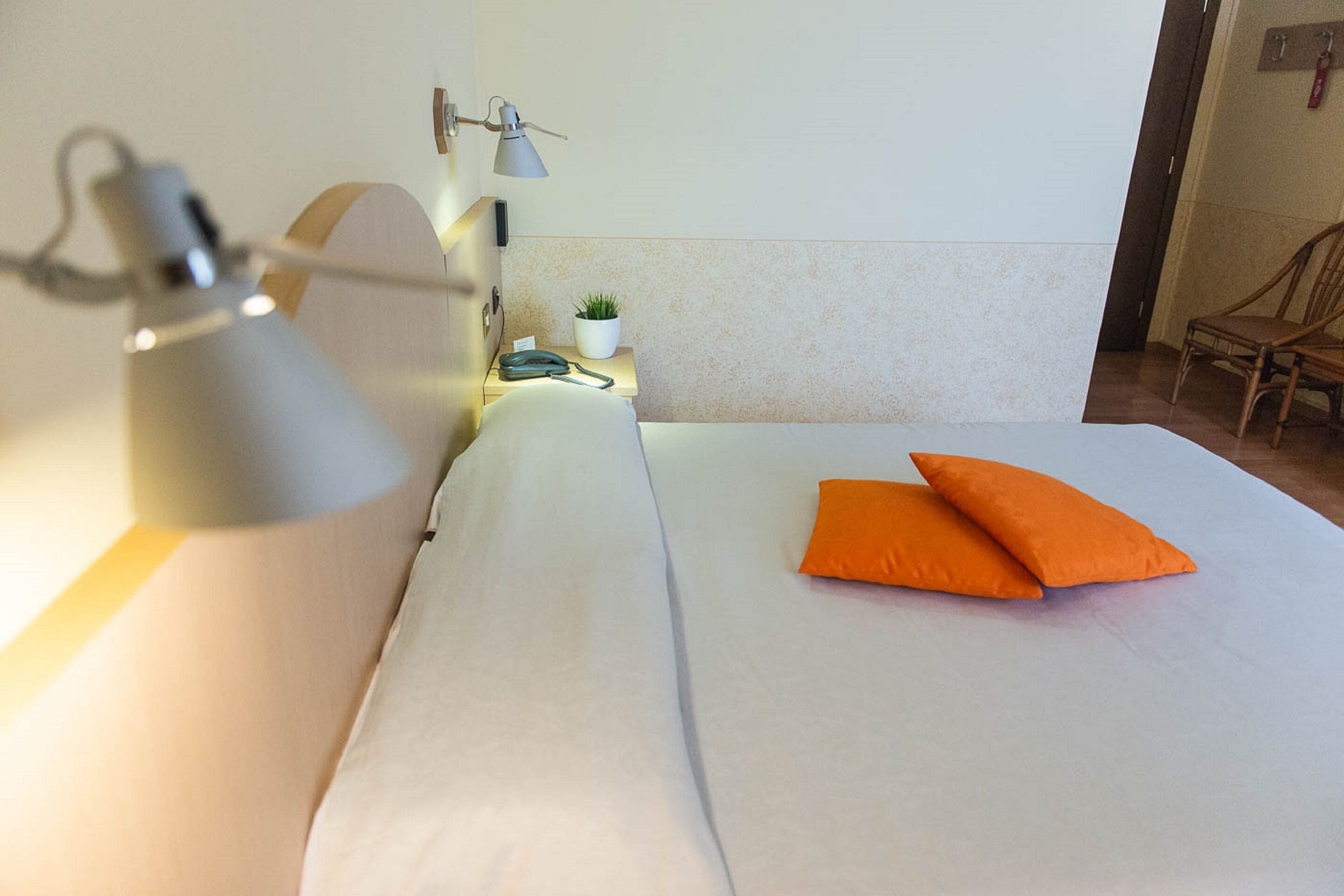 A neatly made bed with white sheets and two orange pillows, a bedside lamp, a small plant, and a chair in the corner.