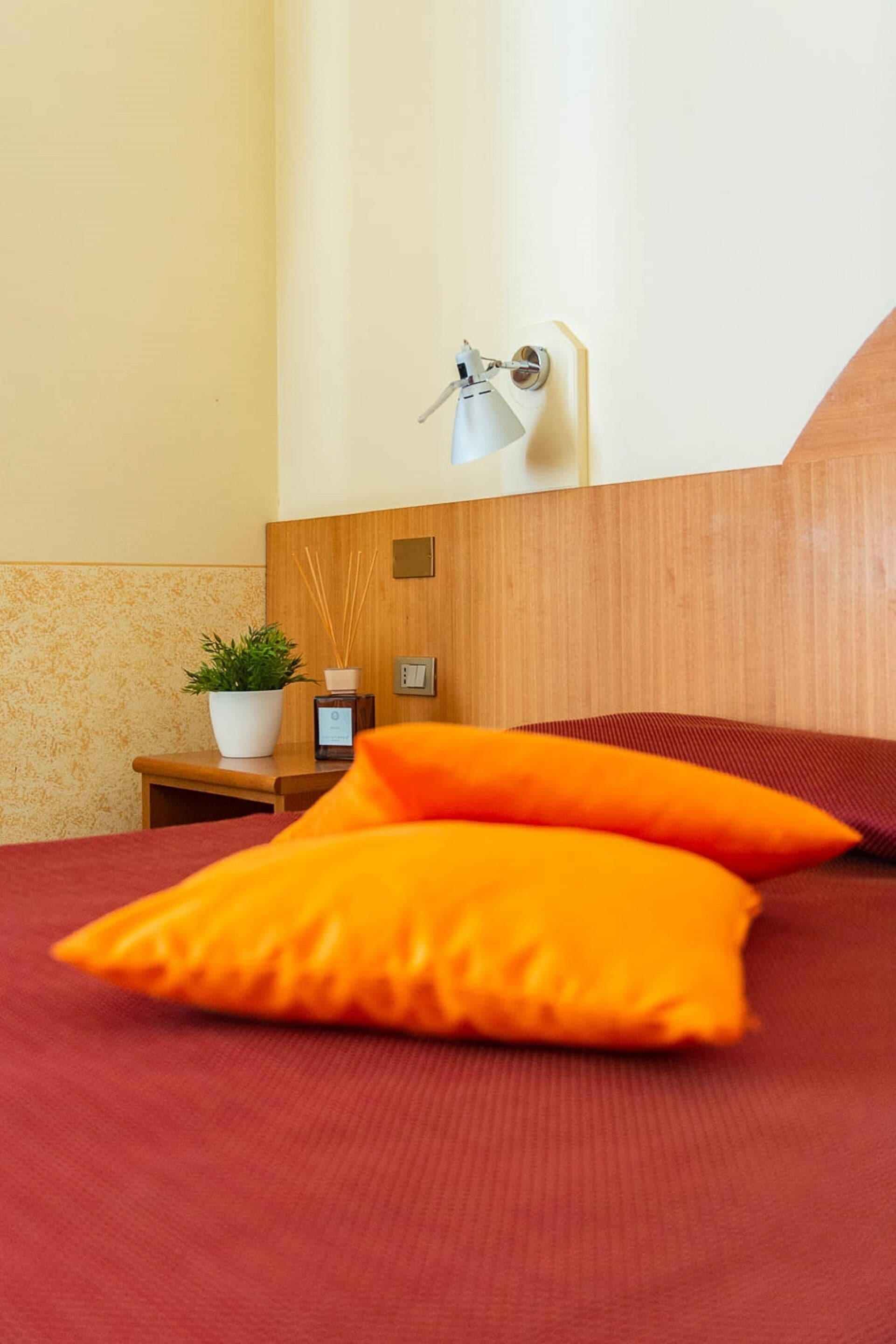 A hotel room with a wooden headboard, a bedside table with a plant, a lamp, and two orange pillows on the bed.