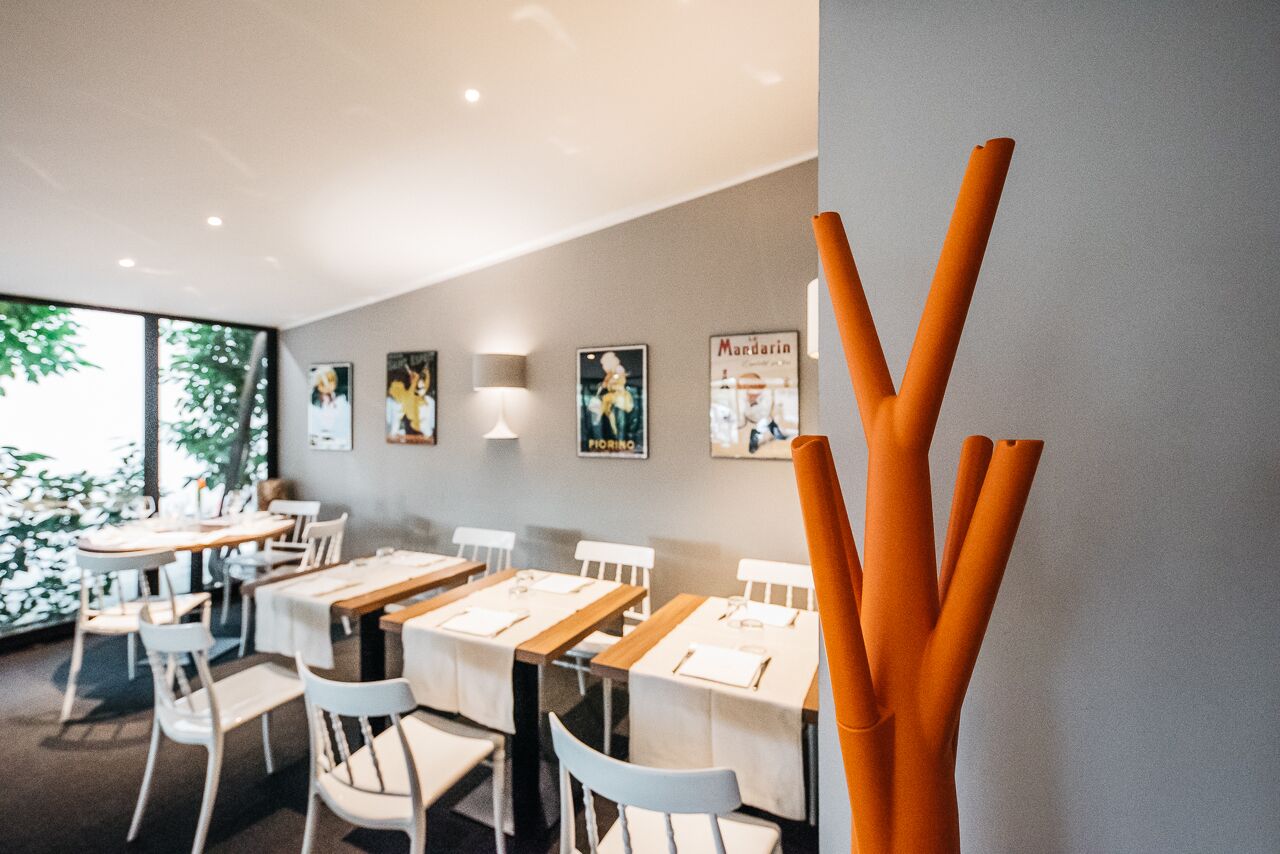 A restaurant interior with tables set for dining, framed pictures on the wall, and a large window with greenery outside.