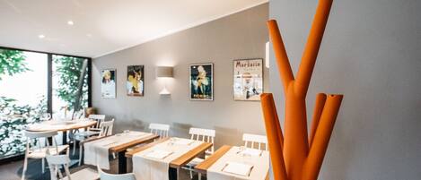 A restaurant interior with tables set for dining, framed pictures on the wall, and a large window with greenery outside.