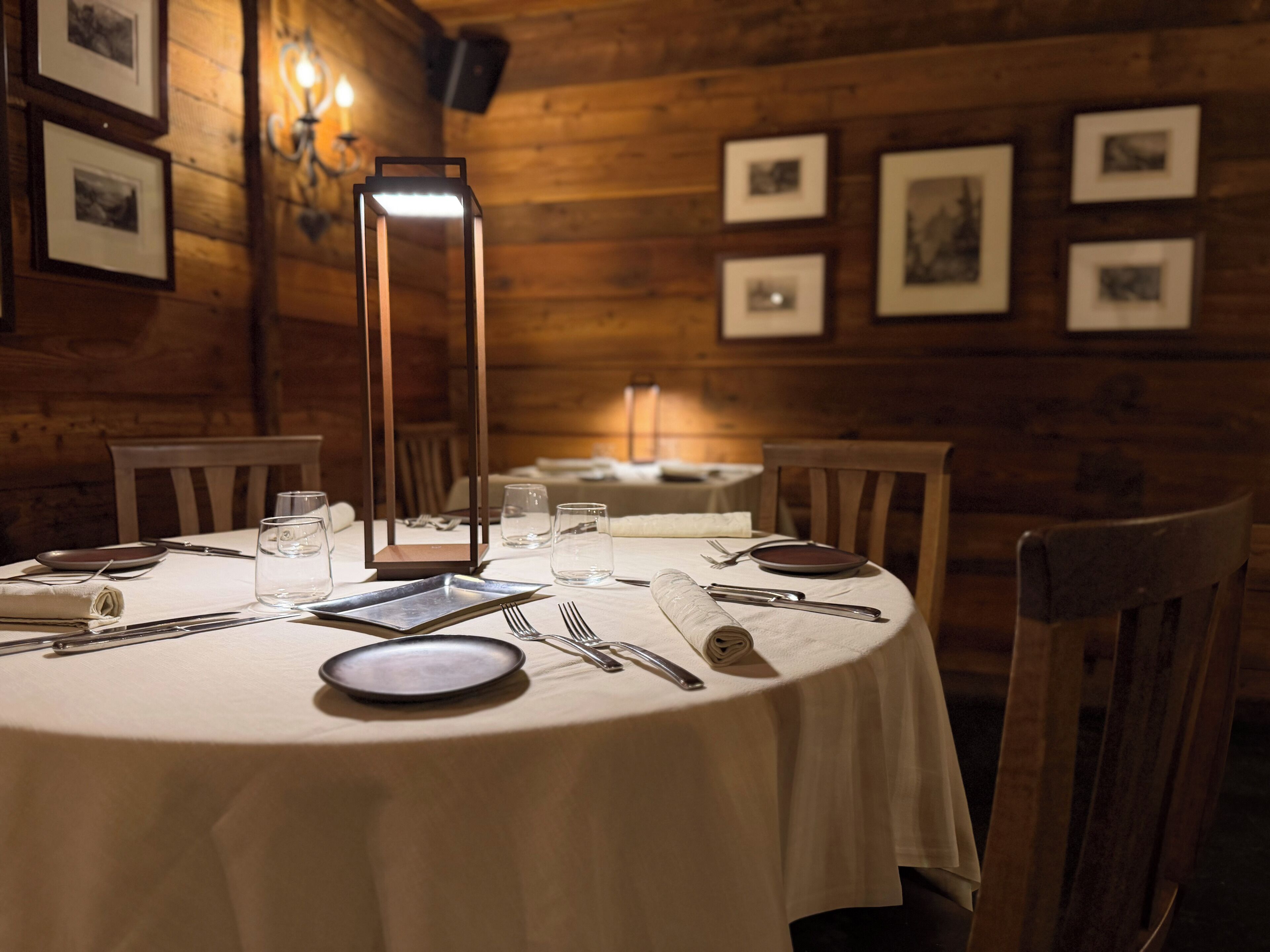 A round dining table with a white tablecloth, surrounded by wooden chairs. A tall, rectangular, illuminated table lamp is centered on the table.