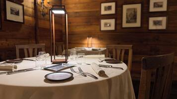 A round dining table with a white tablecloth, surrounded by wooden chairs. A tall, rectangular, illuminated table lamp is centered on the table.