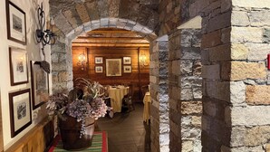 A stone-walled corridor leading to a dining area with wooden beams, framed pictures, and a large potted plant. - Maison Saint Jean (Courmayeur)