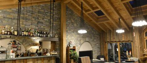 Um interior rústico com área de bar, paredes de pedra, vigas de madeira e luminárias pendentes.