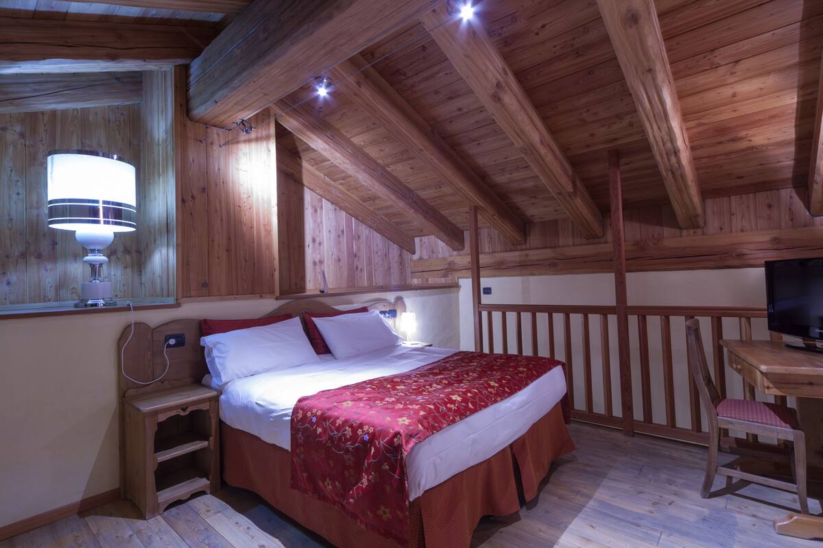 A wooden cabin bedroom with a bed, bedside table, lamp, and a TV on a wooden desk.
