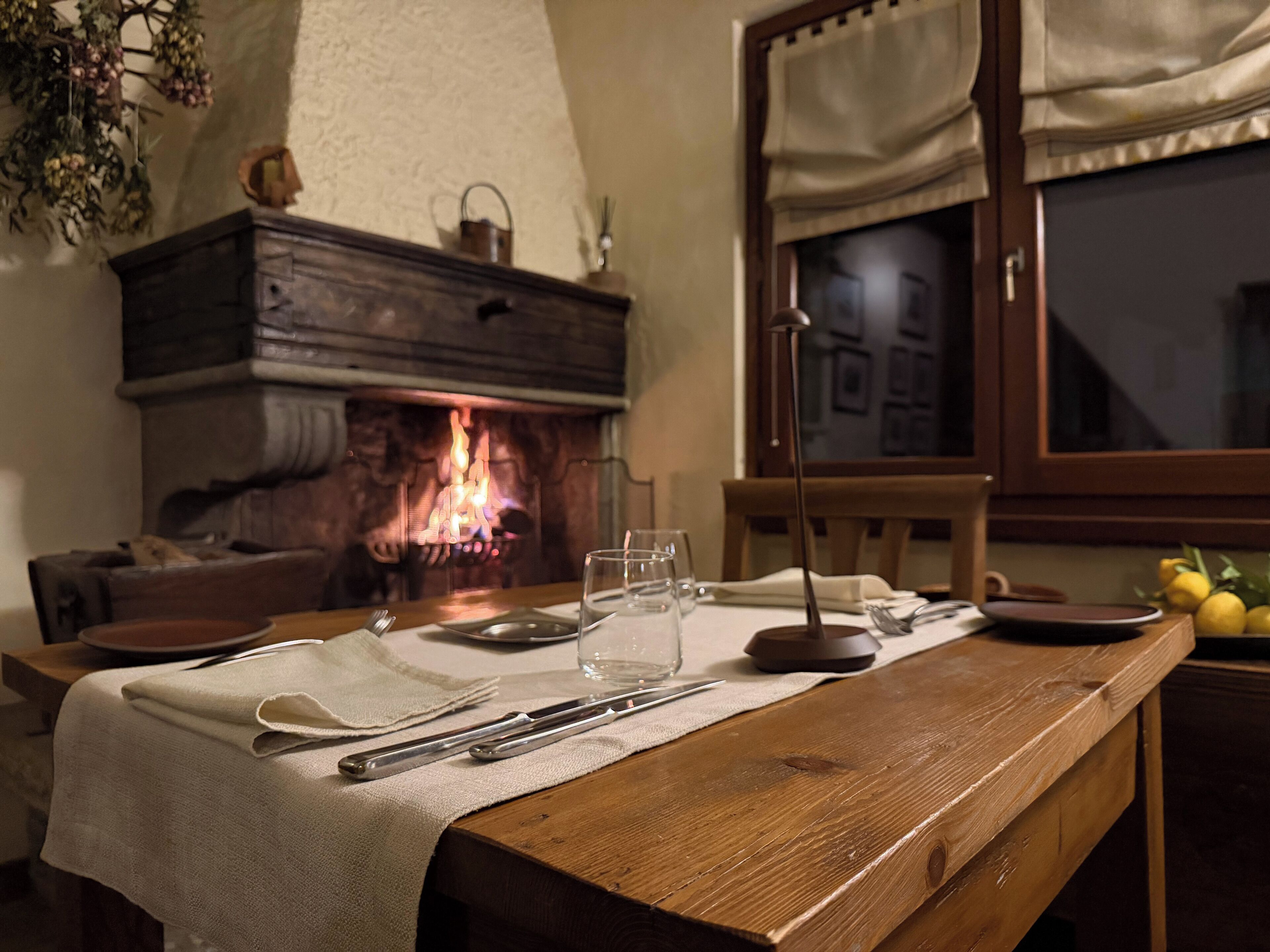 A dining table set with cutlery and glasses, a fireplace with a visible fire, and a window with curtains.