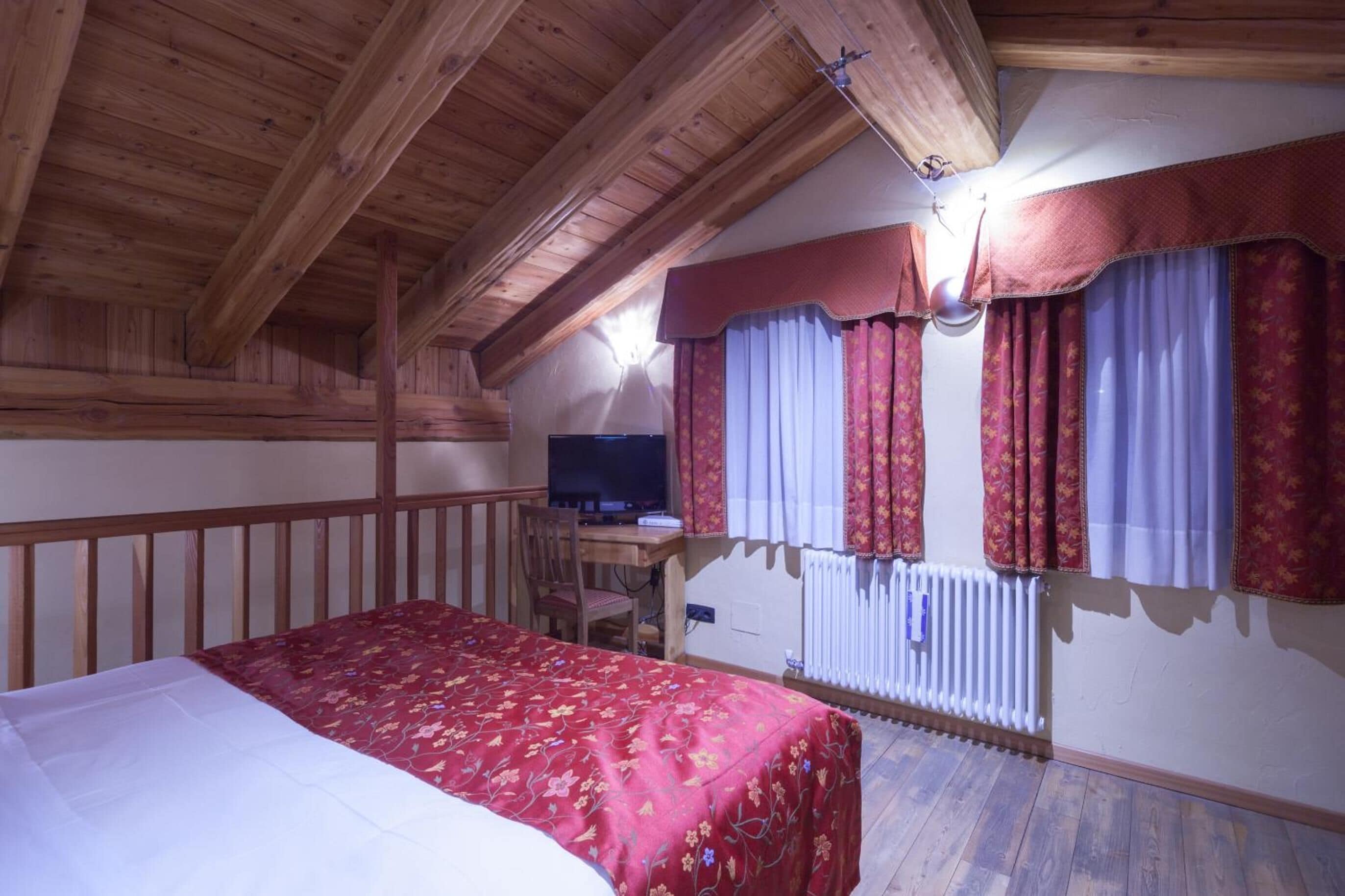 Quarto com teto de madeira, cama com colcha florida, escrivaninha com computador e radiador.