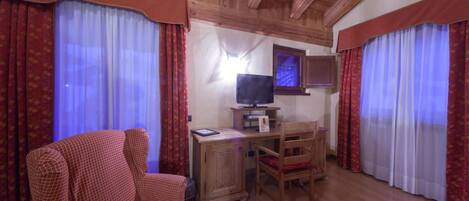 A cozy room with a wooden ceiling, a television on a wooden stand, a desk with a chair, and red curtains.