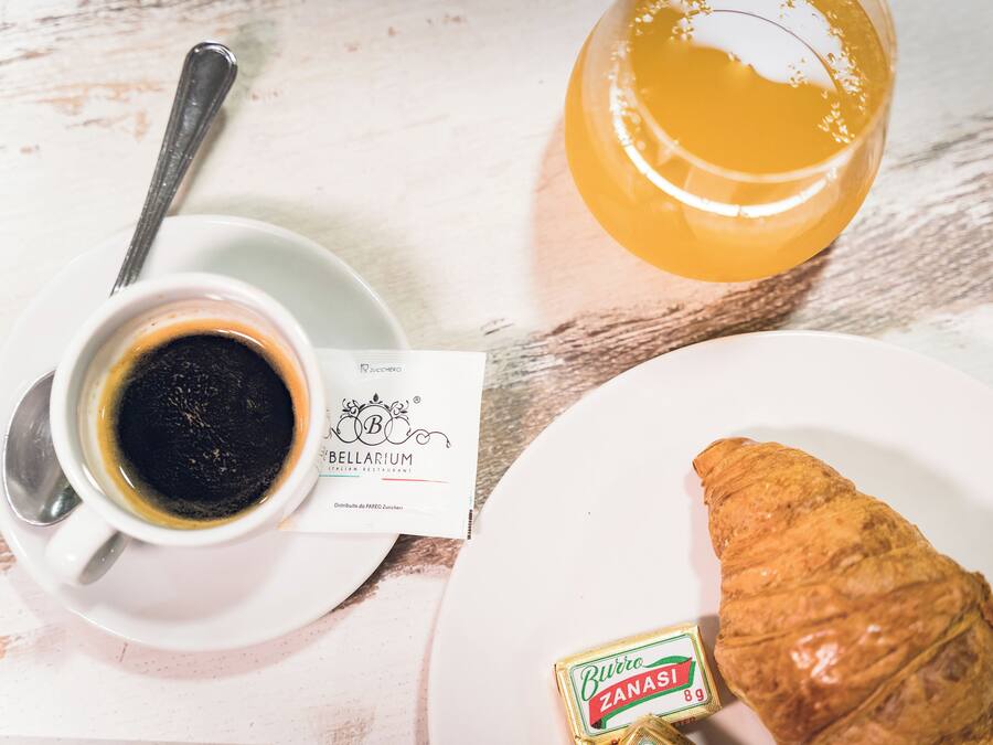 Disposição para o pequeno-almoço com uma chávena de café, um copo de sumo de laranja, um croissant e uma pequena caixa de biscotti sobre uma mesa branca.