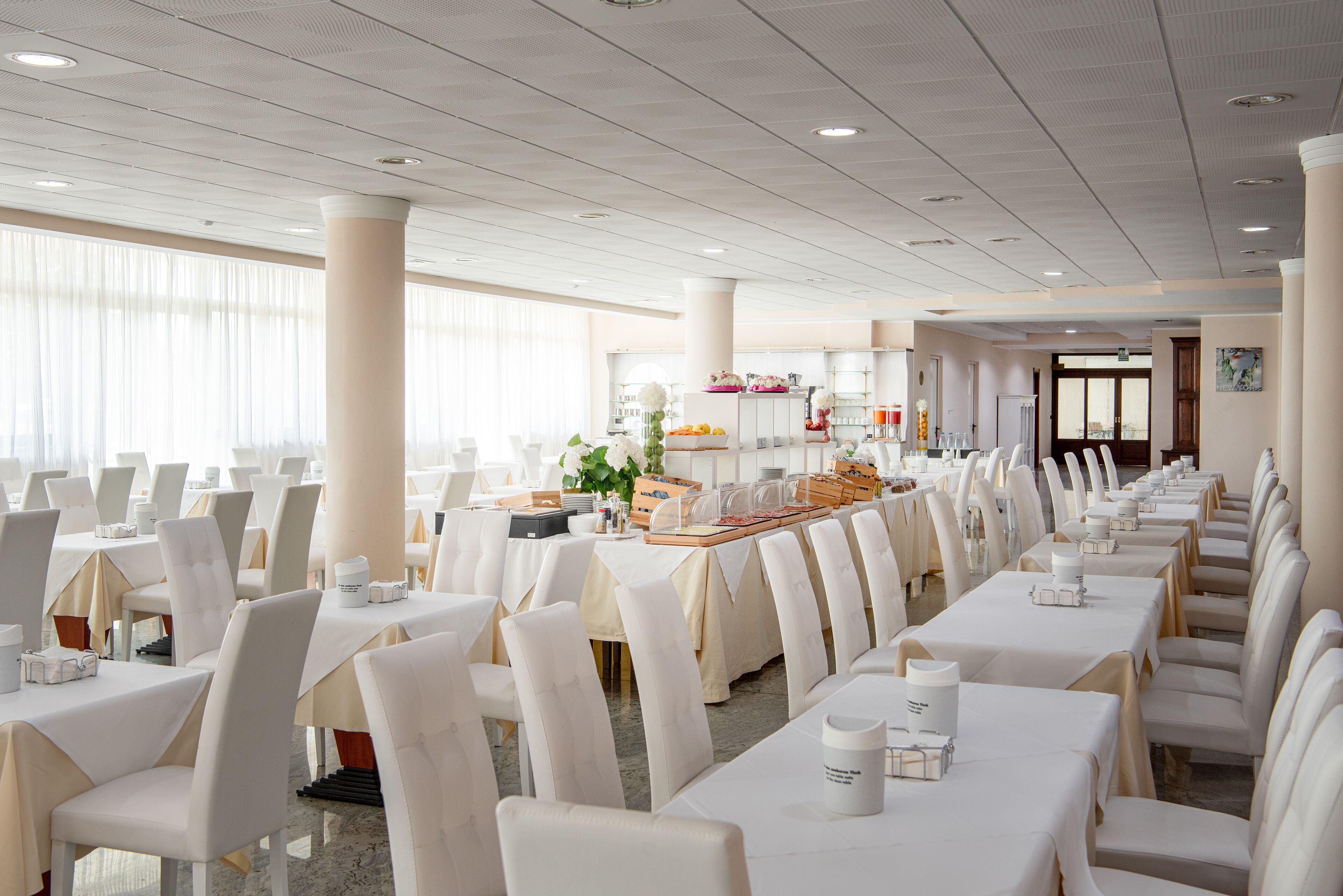 Un amplio salón de banquetes con filas de mesas y sillas blancas, una zona de buffet y grandes ventanales que dejan entrar luz natural.