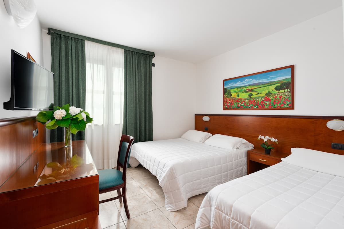 A hotel room with two beds, a desk with a TV, a chair, a painting on the wall, and a window with curtains.
