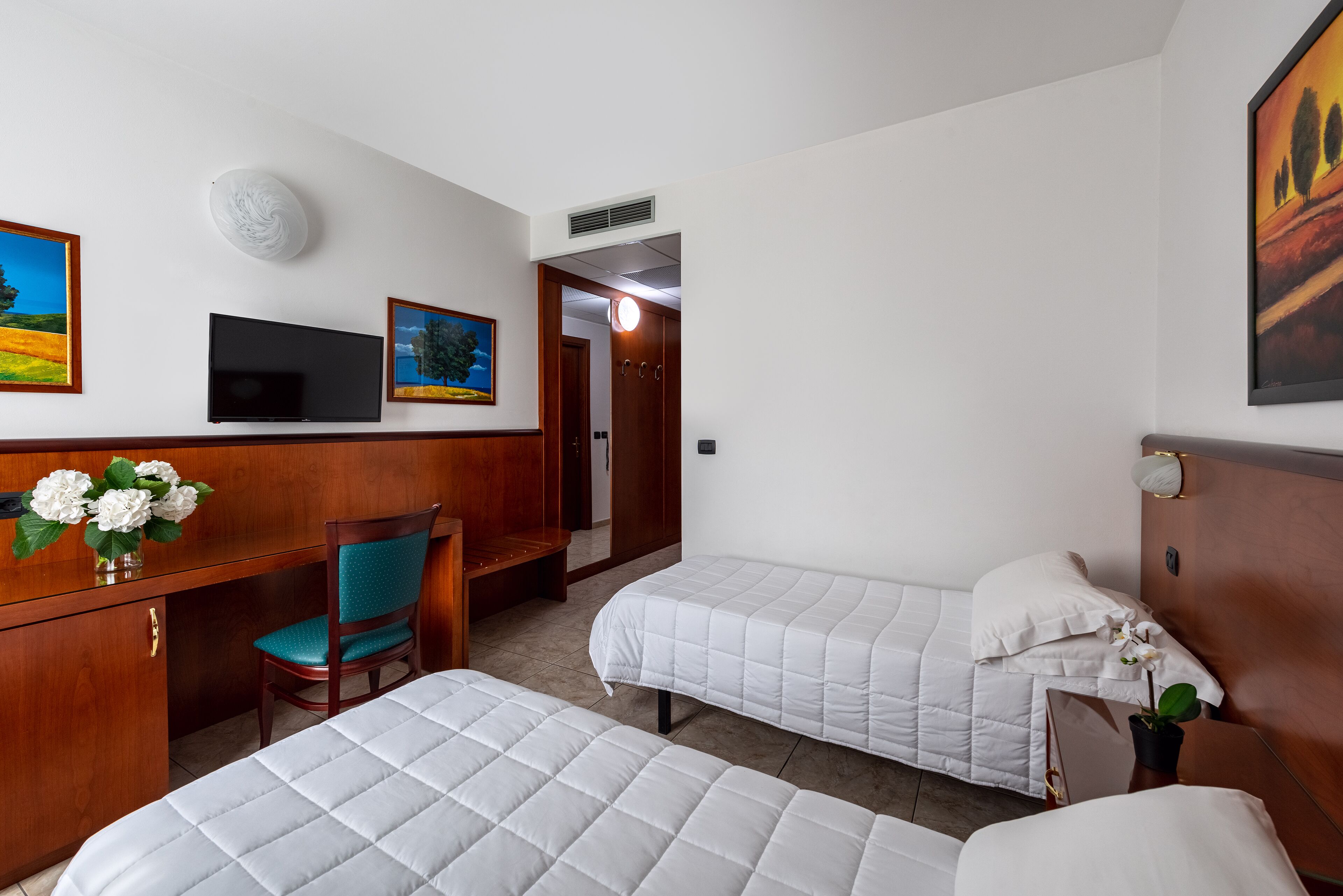 A hotel room with two beds, a desk with a chair, a television, and artwork on the walls.