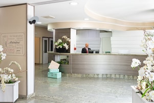 Uma área de recepção de hotel com design moderno, apresentando um balcão de atendimento, um recepcionista e orquídeas brancas decorativas.
