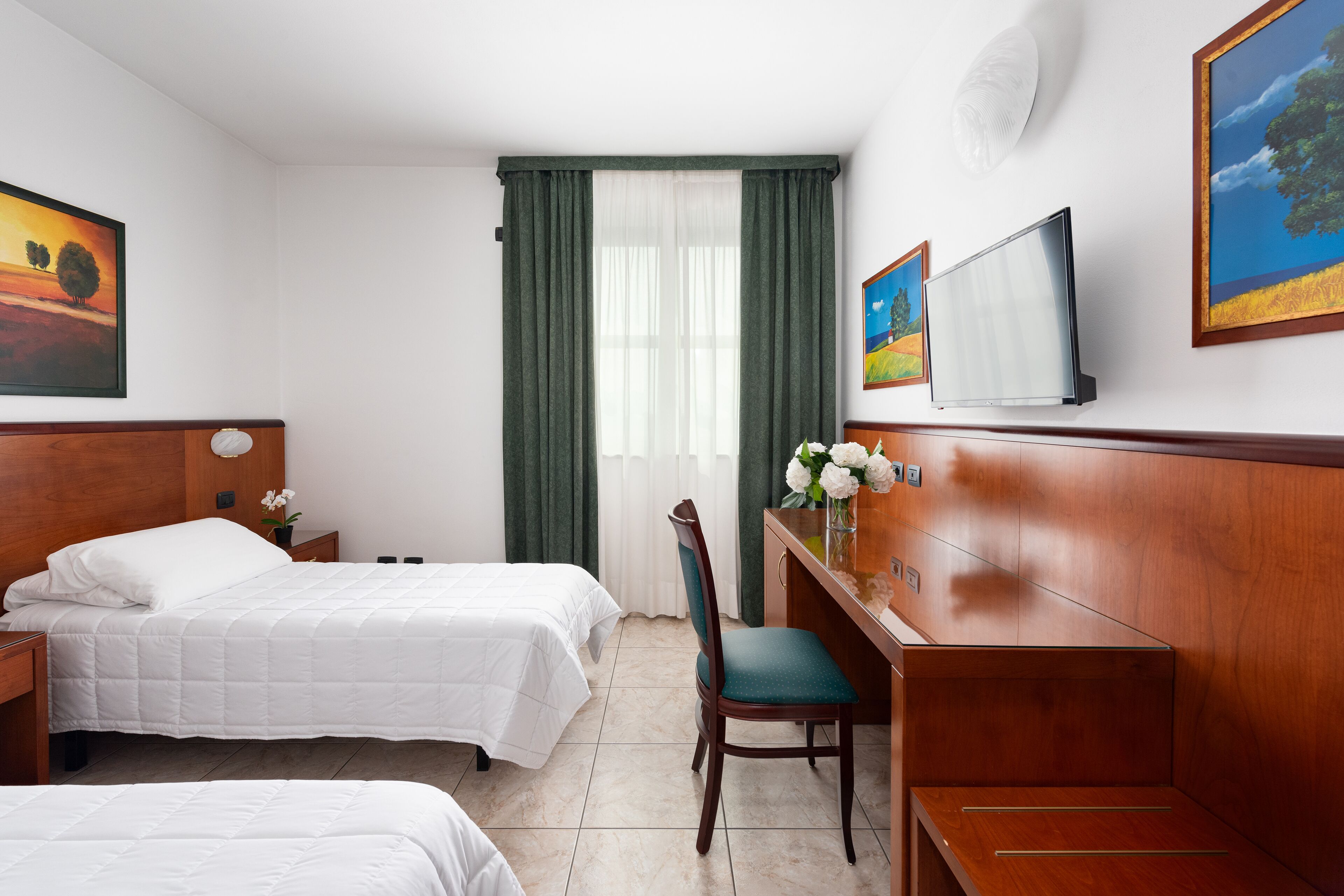 Habitación de hotel con dos camas, un escritorio, una silla, un televisor y obras de arte en las paredes.