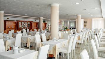 Un'ampia sala da pranzo con diversi tavoli e sedie bianche, caratterizzata da imponenti pilastri e pensili in legno sullo sfondo.