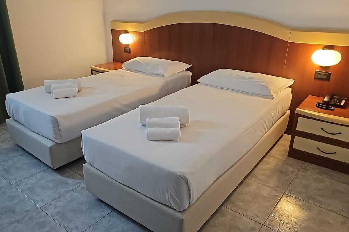 A hotel room with two single beds, each with a pillow and a towel, a wooden headboard, bedside lamps, and a nightstand with a phone.