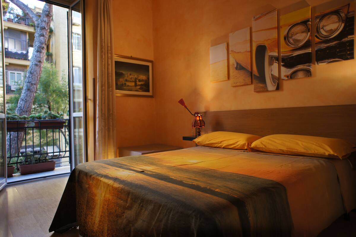 Camera d'albergo con un letto, un comodino, una lampada e una finestra vista su un albero e un balcone.