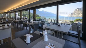 A modern dining area with tables and chairs, large windows offering a view of a lake and mountains. - Riviera (Riva del Garda)