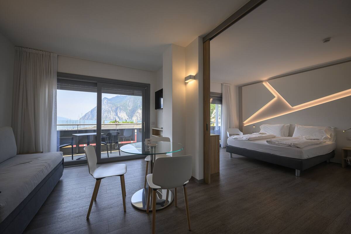Una habitación de hotel moderna con una cama, una mesa de comedor de vidrio y sillas, un balcón con vistas a montañas, y un ventanal grande.